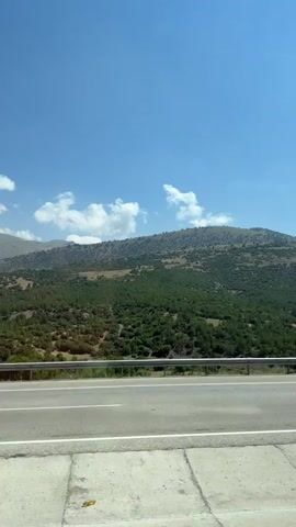 The video captures a view from a moving vehicle, looking out towards a mountainous landscape under a clear blue sky with scattered white clouds. In the foreground, a paved road with white lane markings is visible, separated from the landscape by a metal guardrail. The mountains are covered in dense green vegetation, with some bare, rocky patches at the higher elevations. Several trees of varying sizes are scattered along the lower slopes and near the road. A white car passes by on the road in the latter part of the video. The scene appears to be a scenic drive through a natural environment.