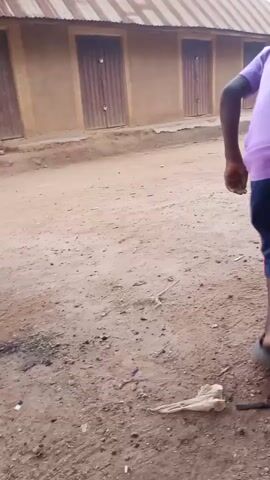 A person is playing with a tire in a dirt area. The person is wearing a purple shirt and dark shorts. They are pushing the tire with a stick. There are buildings in the background.