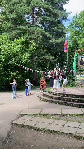 The video opens with a wide shot of an outdoor performance space. In the foreground, a paved area leads to a tiered wooden stage adorned with greenery. A group of children stands on the paved area, facing the stage. On the stage, a group of individuals, likely young people, are engaged in what appears to be a theatrical performance. They are positioned on the top tier of the stage, with some in the process of interacting with a large, wooden, cog-like prop. The backdrop features lush green trees and a clear blue sky with some clouds. Red, white, and green triangular flags are strung across the background, adding a festive atmosphere. A Russian flag and a green banner are also visible. The overall setting suggests a community event or an outdoor play.