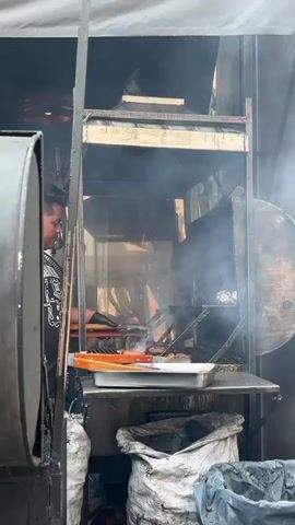 A man wearing a black shirt is grilling meat inside a metal enclosure filled with smoke. The enclosure has a grill with flames burning beneath it. A tray with grilling utensils is on a shelf below the grill. There are sacks of charcoal below the shelf.