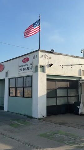The video shows the exterior of a building on a clear day. An American flag waves from a flagpole on the roof of the white building. The building has a sign that says "Cafe" and lists a phone number for phone orders. There are windows on the side of the building and a large garage door. The building appears to be a commercial establishment. The ground in front of the building is paved.