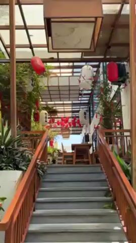 The video shows a low-angle view of a staircase leading up to an outdoor dining area with a Japanese-themed aesthetic. The staircase is made of gray concrete with wooden railings on both sides.  Hanging above the staircase and dining area is a lattice-like structure with square panels, some of which are translucent.  Red and white Japanese lanterns are strung along the structure. Green plants are placed along the sides of the staircase and around the dining area. Tables and chairs are visible in the dining area. The lighting suggests it is daytime. There is music playing.