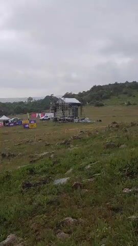 This media file captures an outdoor event, likely a festival or market, set in a rural landscape. The scene is dominated by a large open field dotted with numerous white tents, suggesting vendor stalls or activity zones. A scaffolding structure with a roof is visible in the background, possibly a stage or an information booth. A yellow crane is also present, indicating ongoing setup or construction for the event. Several vehicles, including white cars and a truck, are parked amongst the tents. The ground is covered in grass and rocks, typical of a natural, undeveloped area. In the far distance, rolling hills and a hazy sky suggest a mountainous or hilly terrain. The weather appears overcast, with a cloudy sky. The overall mood is one of preparation and anticipation, as the event is clearly in its early stages or just beginning. While not explicitly stated, the presence of tents and a stage hints at a gathering of people. The text "HEYLE" is visible on a banner. The location is identified as Antarut, Armenia.