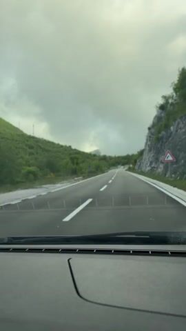 From the perspective of the driver, a car travels down a paved road with white lane markings. Two other cars are visible ahead, approaching on the same road. The landscape is mountainous, with green vegetation covering the slopes on either side. A cliff face is visible on the right. The sky is overcast with a heavy cloud cover. The dashboard of the car from which the video is taken is visible in the foreground.