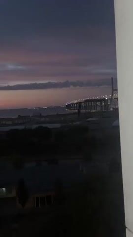 The video shows a time-lapse of a sunset over a city. The sky transitions from a dark blue to shades of pink and orange as the sun sets. In the foreground, there are silhouetted buildings and trees. In the background, a large bridge is visible, illuminated by lights. The overall scene is one of a city preparing for nightfall.