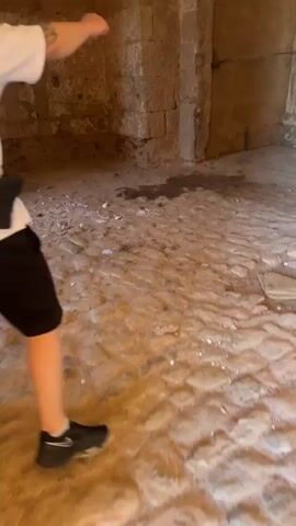 The video shows a man walking towards a large wooden door in a stone room. The room has a stone floor and walls. The door is weathered and old. There is some light coming through gaps in the door. The man is wearing a white shirt, black shorts, and a black bag over his shoulder. The camera follows the man as he walks towards the door.