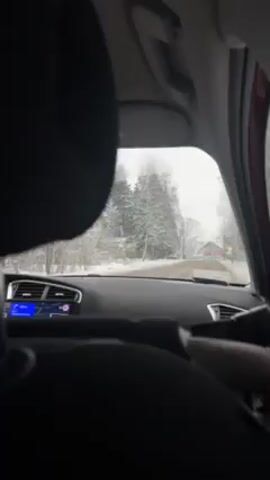 The video shows a car driving down a road covered in snow. The view is from inside the car, looking out the windshield. Snow-covered trees and a red building can be seen on the sides of the road. The car's dashboard and GPS navigation system are visible. It appears to be daytime with overcast sky.