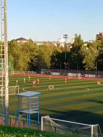 The video shows a soccer field with several players practicing. The field is artificial turf and surrounded by a fence. There are two goals visible on the field, and a small building with a blue roof and glass windows, possibly a referee's booth, is situated near the sideline. In the background, there are trees and buildings. The sky is clear and blue, indicating daytime. The lighting suggests it is either early morning or late afternoon, with the sun casting long shadows. Some players are wearing red jerseys, while others are in yellow. They appear to be engaged in drills, with some running and others kicking the ball. A tall metal structure, possibly a light pole or scoreboard support, is prominent in the foreground on the right side of the image.