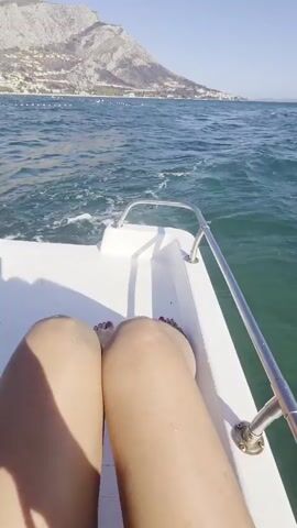 A first-person perspective video shows legs and a small black dog with a blue collar on a white boat moving through the water. The dog is being petted by a person whose hand is visible. In the background, a coastline with buildings and a mountain is visible under a clear sky. The water is choppy and blue, with white foam trailing behind the boat. The overall atmosphere is that of a leisurely boat trip on a sunny day.