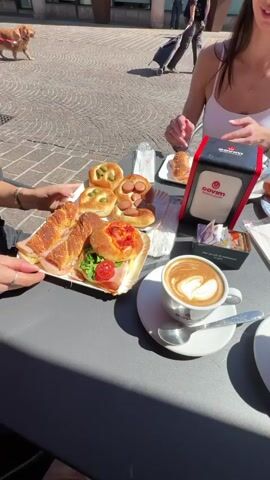 The video shows a group of women enjoying a meal at an outdoor cafe in Italy.  They are seated around a black table that holds an assortment of pastries, sandwiches, and cups of coffee with latte art.  Bottled water and napkins are also present on the table. One woman, wearing a white tank top and sunglasses, uses her phone, while another woman next to her looks down at her phone. A dog on a leash is visible in the background. The area outside the cafe is paved with red bricks, and other people are seen walking by, some pulling luggage. The weather appears sunny and bright. Buildings are visible in the background, one with "SANPAOLO" inscribed on it.