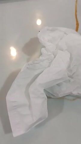 A crumpled white tissue paper lies on a light-colored, smooth surface. Streaks of a brown substance are visible on the surface, trailing from behind the tissue. There are two bright, oval reflections on the surface, suggesting a light source above. A portion of a red and yellow sign with text is visible in the upper right corner of the frame. The video is shot from a top-down perspective.