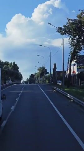 This is a vertical, first-person perspective video of a road on a sunny day. The camera is positioned on a moving vehicle. The road is asphalt with a white line marking the right lane. On the left side of the road are trees and a sidewalk. On the right side of the road are more trees, streetlights, and buildings. The sky is blue with a large, fluffy white cloud. There are other vehicles on the road, including a white truck in the foreground on the left and a truck in the distance on the road. The overall impression is of a typical suburban or rural road during the daytime.