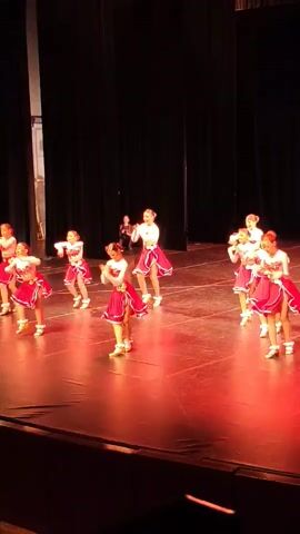 A group of young girls in matching red and white dance costumes with short, ruffled skirts and white tops are performing on a stage. They are wearing white roller skates. The stage is lit with red lighting. They are arranged in a semi-circle, with some girls in front and others behind. They are all facing the same direction and performing synchronized dance moves, including arm movements and leg kicks. The background is a dark curtain. The girls appear to be performing for an audience, although no audience members are visible in the frame. The video is short and captures a portion of their performance.