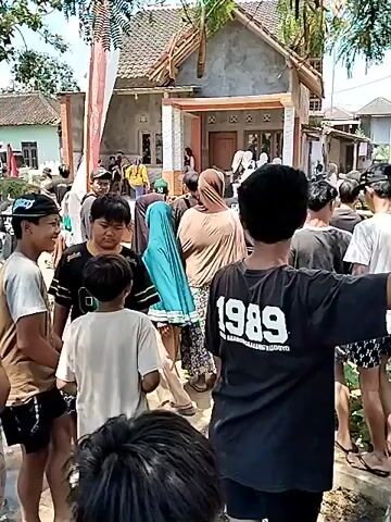 The video begins with a close-up of green, elongated pods hanging from a tree. It then pans to show a group of young men gathered outdoors in what appears to be a residential area. One of the men is wearing a black t-shirt with "1989" printed on the back. There are houses in the background, and some people are standing on a porch. The scene suggests a casual gathering or possibly an event.
