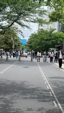 The video shows a street scene, likely on a university campus or in a similar pedestrian-heavy area. A crowd of people, mostly young adults and teenagers presumably students, is moving in both directions along the paved street. Some are walking, while others are riding bicycles and a scooter. The street is lined with bollards and white dashed lines, suggesting it is a pedestrian zone or has restricted vehicle access. Trees are visible on either side of the street, offering some shade, and buildings are in the background. There is some construction visible in the distance on the left side of the frame. The weather appears to be overcast, and the lighting is somewhat flat.