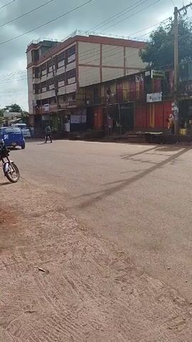 The video shows a street scene in a small town. A red motorcycle is parked on the side of the road next to a pile of rubble that is partly covered by orange netting. A sign for "Miracle Revival Fellowship Pentecostal Church- Gachie Branch" is visible above the sidewalk. The road itself is mostly empty, with a few pedestrians and vehicles visible in the distance. The buildings along the street appear to be a mix of residential and commercial properties, and the overall environment seems somewhat run-down. The sky is overcast, and power lines are strung across the street.