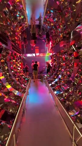 The video shows a person walking down a hallway. The hallway walls and ceiling are covered in mirrored surfaces and colorful, jagged shapes, resembling a kaleidoscope. Several other people are visible in the hallway ahead, and some reflections of people can be seen in the mirrored surfaces. The floor of the hallway is a smooth, light-colored surface with a colored light projection on it. The overall impression is that of an immersive art installation.