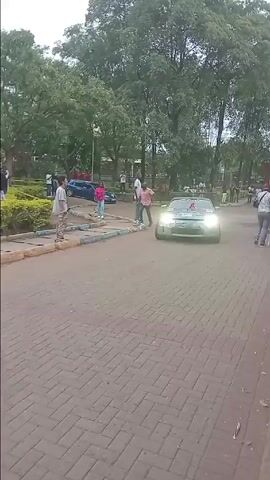 The video shows a silver Nissan 350Z with a damaged hood driving on a brick road. The car has stickers on it and a rack on the back. The road is surrounded by bushes and trees, and a crowd of people is standing along the edge of the road. The camera follows the car as it drives around the corner.