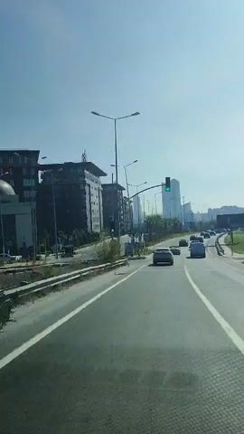 The video shows a view from a vehicle traveling on a highway. The highway has multiple lanes and is flanked by a concrete barrier and a grassy embankment. In the distance, several modern buildings are visible, including a mosque with a dome and minaret on the left side of the road. Traffic lights are present, some of which are green. There are cars on the highway, and the sky is clear and blue. A billboard is visible on the right side of the road. The overall impression is of a modern urban or suburban environment on a sunny day.