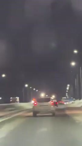 The video depicts a nighttime scene on a multi-lane highway, likely in a cold climate given the presence of snow along the sides of the road. The camera appears to be mounted inside a vehicle that is following a light-colored SUV. Numerous streetlights illuminate the road. In the distance, a vehicle with flashing red lights, possibly an emergency vehicle, is visible. The video seems to be taken through a wet windshield, as there are water droplets visible on the glass.