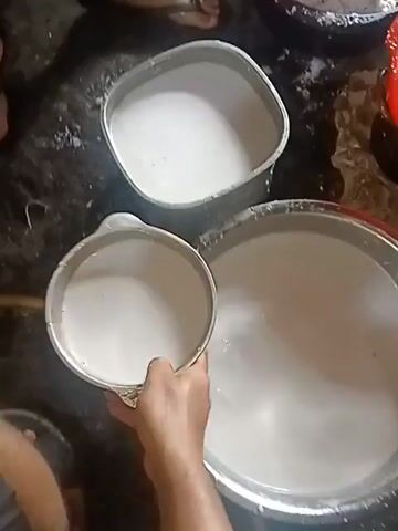 A high-angle medium shot captures a cooking process in a rustic environment. In the foreground, a large, shallow metal pan rests on a dark, possibly stone or concrete surface. The pan contains a reddish-brown mixture that appears to be simmering or boiling, with hints of oil visible. A silver spatula or ladle is positioned within the pan, resting on the right side of the mixture.

To the right of the pan, a person's hands are visible as they pour a white liquid, likely coconut milk or cream, from a clear plastic cup into the cooking mixture. The liquid is cascading into the pan, creating ripples and swirls on the surface of the reddish-brown base.

In the background, two metal containers are partially visible. The one on the left is a square-shaped metal bowl containing a creamy white substance, possibly uncooked ingredients or a prepared component. The other container, further back and to the right, is out of focus but appears to be round and metallic.

The overall environment suggests a kitchen or outdoor cooking area, with a rough, textured surface beneath the cooking equipment. The lighting is dim, typical of indoor or shaded outdoor settings. The focus is primarily on the action of pouring the liquid into the pan, highlighting the preparation of a dish.