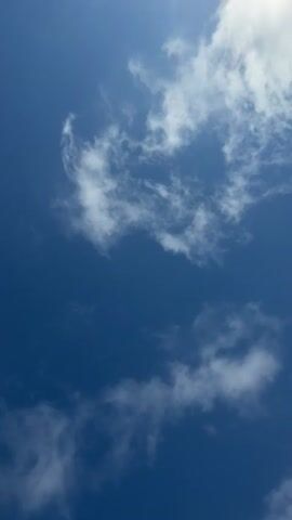 The video shows a close-up, upward-facing view of a bright blue sky with scattered white clouds. The clouds are wispy and irregular in shape, with some appearing more dense and others more translucent. The sunlight is strong, creating a bright exposure and some lens flare in the upper left corner. The perspective is tilted, giving the impression of looking straight up from the ground. There are no people, objects, or discernible actions visible in the video, only the natural elements of the sky.