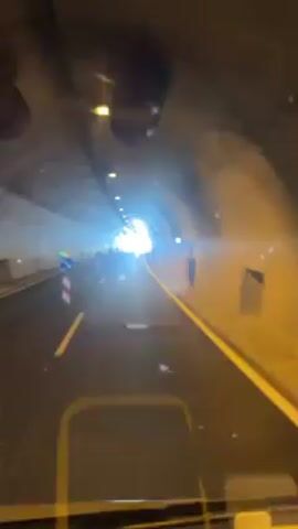 The video shows a car driving through a tunnel. There are road cones along the left side of the road. The light at the end of the tunnel is very bright. There are round black ventilation units on the ceiling. The road is black, with a yellow stripe to the right. The walls of the tunnel are concrete and smooth. There are yellow road lines in the middle of the lanes.
