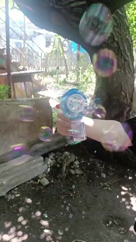 A close-up shot shows a person's hand holding a blue and white bubble gun. The bubble gun is emitting numerous soap bubbles that float upwards and around the frame. The background is outdoors, with dirt and debris on the ground, rocks, a tree trunk, and some distant structures. The lighting suggests it is daytime. The person's arm is visible, wearing a light-colored sleeve.