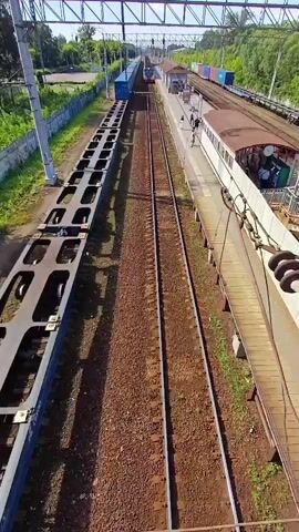 The video is an aerial shot of a train station with two sets of train tracks. On the left track, there is a long, flatbed train car with multiple empty platforms. On the right track, there is a passenger train stopped at a station platform. Several blue shipping containers are visible on the tracks in the distance. A wooden ramp leads from the platform to a higher walkway. The station is surrounded by greenery and trees.
