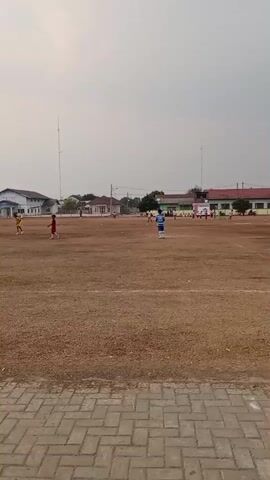 The video shows a wide shot of an outdoor soccer field under a hazy sky. The field is covered in dry, brown grass, with white lines marking the boundaries. Several young people, presumably boys, are scattered across the field, wearing brightly colored jerseys and shorts, and appear to be playing soccer. One boy in a blue and white striped jersey is standing in the middle of the field, facing towards the left. In the background, there are several buildings, including a long white building with a blue roof and a red-roofed building. Two tall antenna towers are visible in the distance. The foreground of the video shows a paved walkway made of brick. The overall atmosphere suggests a casual game or practice session taking place on a warm day.