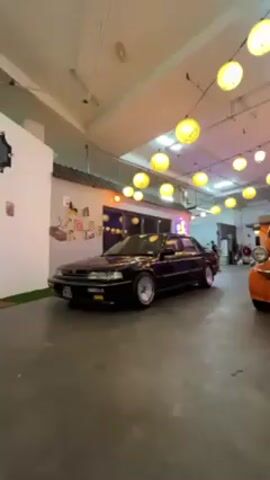 The video shows a black Honda Accord parked indoors. The car is the main focus, shown from the front quarter view. It features a modified suspension, bringing it closer to the ground, and aftermarket wheels. The environment is brightly lit with numerous string lights that resemble lanterns. To the left, there's a wall decorated with photos and drawings, giving it a personalized or cafe-like ambiance. An orange vehicle is partially visible on the right edge of the frame, suggesting a collection of vehicles in the space. A person can be seen in the background moving. The floor is a concrete surface.