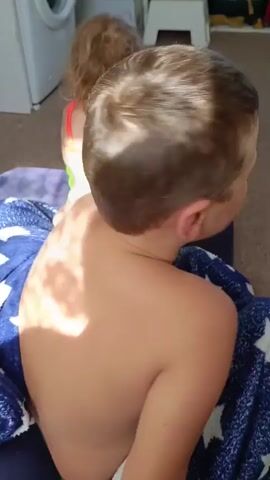 A close-up shot focuses on a young boy sitting down and wrapped in a blue blanket with white stars. The boy is looking directly at the camera with a smiling expression. His eyes are partially closed, and his cheeks are flushed. Sunlight illuminates his face, casting dappled shadows. In the background, another child, a girl, is visible, also wearing a blanket. A washing machine is partially visible to the left of the frame. The setting appears to be indoors, with a floor that looks like carpet or tile.