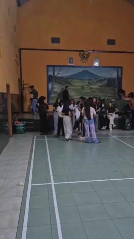 The video captures an indoor scene, possibly a gymnasium or community hall, with a large mural of a mountain landscape on the far wall. The walls are painted a bright orange. The floor is tiled in a green and white grid pattern, marked with white lines indicative of a court, possibly for badminton. In the center of the court, several young people are gathered. Two women, one in a white and patterned skirt and the other in patterned pants, appear to be dancing or posing. A group of onlookers stands further back, some sitting on benches or chairs against the mural wall. To the right side of the court, a drum set and other musical equipment are visible, suggesting a performance or band setup. The lighting is moderate, with overhead lights illuminating the space. The overall atmosphere appears to be casual and social, possibly a gathering or event.