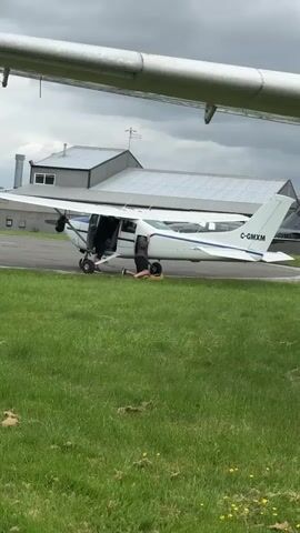 The video shows a small white and blue Cessna aircraft on a tarmac with its door open. A person is kneeling down on the ground, apparently working on the nose wheel of the plane. Another person is standing in the doorway of the aircraft, looking on. The aircraft is parked on the edge of a grassy area. In the background, there is a building with a silver roof. Another aircraft is partially visible on the far right side of the frame. The video is taken from under the wing of another plane. The weather appears overcast. The plane's registration number is C-GMXM.