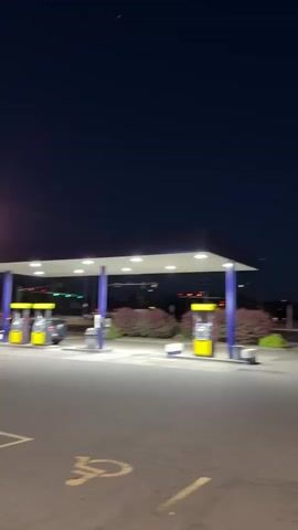 This media file captures a gas station scene at dusk. The setting is an outdoor gas station with a covered canopy sheltering multiple gas pumps. The pumps are distinct with their bright yellow casings and blue support columns. The ground is asphalt, marked with yellow lines, including a visible disabled parking symbol. In the background, there are some businesses with illuminated signs, trees, and other vehicles, suggesting a suburban or commercial area.

The time of day is clearly evening, as indicated by the dark blue sky and the artificial lighting from the gas station canopy and streetlights. The weather appears calm, with no visible rain or strong winds.

The activity depicted is the general operation of a gas station. While there are no explicit actions of fueling a car, the presence of pumps and the overall environment point to this purpose. A lone individual is briefly visible walking in the vicinity of the gas station in some frames, adding a human element to the scene.

Notable details include the motion blur in some frames, suggesting the camera or the surrounding environment is in motion. The lighting creates a slightly surreal atmosphere due to the contrast between the dark sky and the illuminated station. The location cues, such as the architecture and businesses, are typical of a roadside commercial area in the United States. The overall mood is quiet and perhaps a bit lonely, characteristic of a gas station at closing time or in the late evening. There is no discernible text in the visible portion of the media.