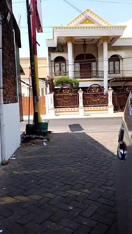 This is a video from a street view, showing a large, ornate white house with gold trim and columns. A brown, decorative metal gate is in front of the house. A street runs in front of the property. On the left side of the street, there is a brick building and a sidewalk paved with dark gray bricks. Two people on a motorcycle are seen riding past the house. Another person on a motorcycle is seen riding past the house later in the clip.