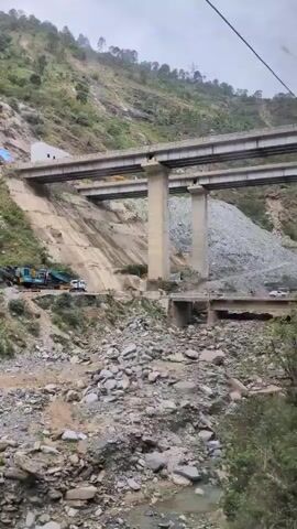 The video shows a mountainous landscape with construction activity. In the foreground, there is a pile of rocks and debris. A dirt road runs along the base of the mountain, with several vehicles, including a truck and construction equipment, visible. Further up the mountain, a large concrete bridge is under construction, supported by massive pillars. The bridge spans across a steep slope where another section of road is visible, with more vehicles. The slopes are covered in vegetation and rock formations. The sky is overcast.