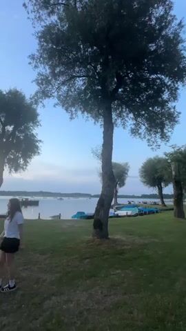 The video opens with a wide shot of a grassy area by a lake at dusk. In the foreground, a young woman stands facing away from the camera, looking towards the water. She is wearing a white t-shirt, black shorts, and white sneakers. Several trees are scattered across the lawn. In the middle ground, a group of people are gathered near the water's edge. Boats are moored along the shore, some covered with blue tarps. The lake is calm, with a few boats visible in the distance. The sky is a clear blue with hints of pink and orange from the setting sun. The video continues with slight panning and zooming, shifting focus between the woman in the foreground and the activities happening by the lake. The overall atmosphere is peaceful and relaxed, suggesting a casual evening gathering.