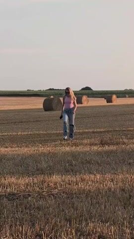 A woman with blonde hair wearing a pink top and ripped blue jeans walks across a dry, harvested field. There are several large round hay bales scattered throughout the field. The sky is clear and pale blue, with a hint of orange near the horizon, suggesting it is either sunrise or sunset.