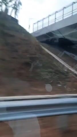The video pans across a scene that appears to be the aftermath of a vehicle accident. The camera is positioned at a low angle, possibly from within another vehicle. The initial frames show a gray structure and debris scattered on the ground. As the video progresses, the focus shifts to a damaged vehicle, possibly a truck, overturned on a dirt embankment. Trees and foliage are visible in the background, suggesting an outdoor, possibly rural or wooded, setting. The lighting indicates it may be daytime, but the sky appears overcast. There are no clearly visible people in the video.