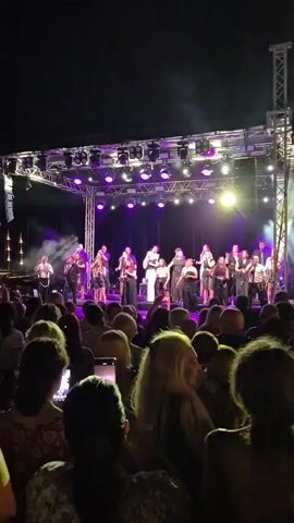 A group of performers stand on a brightly lit outdoor stage. The stage is adorned with purple lights and a large screen or backdrop. The performers are dressed in a variety of outfits, some in formal wear and others in what appears to be traditional or cultural attire. Some are holding instruments, and one individual in the center appears to be speaking or singing into a microphone. Below the stage, a large audience is gathered, silhouetted against the stage lights. Many audience members are holding up their phones, presumably to record the event. The overall atmosphere suggests an outdoor concert or performance taking place at night.