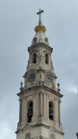 The video shows a close-up of a tall, ornate clock tower against a cloudy sky. The tower is made of light-colored stone and features multiple levels. It has arched openings with bells visible inside. There are two clocks with turquoise faces. The top of the tower is adorned with a golden crown and a cross. Religious chanting or singing is heard in the background.