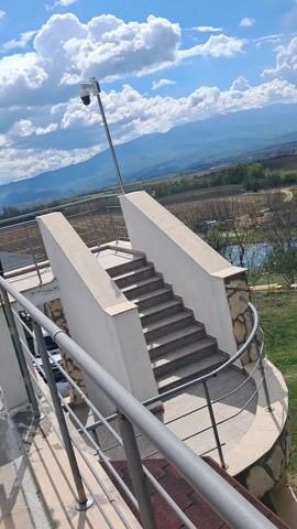 Here's a detailed description of the video:

The video starts with a view from a structure that has steps leading up to it, with railings on both sides. There's a security camera on a pole. In the background are mountains and a bright, cloudy sky. The camera pans to show a green, grassy area below. People are sitting at wooden picnic tables and on the grass. There is also a small pond. The camera continues to pan, revealing more picnic tables, trees and a wooden structure. The video ends returning back to its original position.