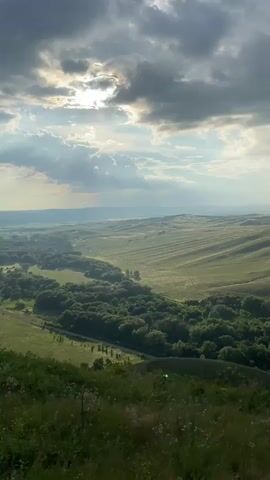 A high-angle video shows a vast green landscape under a dramatic sky. The sun peeks through dark, cloudy formations, casting rays of light onto rolling hills and a valley below. A winding river or road is bordered by dense green trees, creating a stark contrast with the open, lighter green fields that stretch into the distance. The foreground is dominated by blurry green grass and wildflowers. The overall impression is one of natural beauty and a sense of great expanse.