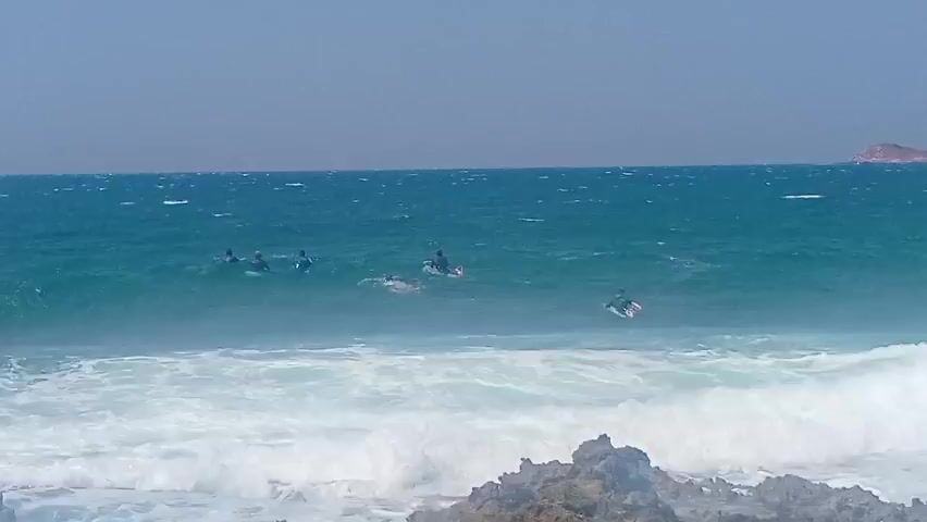 This is a video of a group of surfers in the ocean. The water is turquoise and choppy with white foam from waves crashing. There are several surfers spread out in the water, some riding waves, others paddling. In the foreground, there are dark, rocky shorelines. The sky is clear and blue. The overall impression is a sunny day at the beach with surfers enjoying the waves.