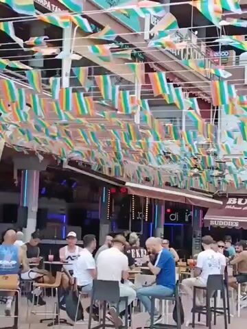 A video shows a street lined with shops, cafes, and restaurants, decorated with numerous colorful rainbow flags strung overhead. People are seated at outdoor tables, eating, drinking, and conversing. The buildings have awnings and various signs. Some establishments include "BUDDIES", "BAR MEICKER", and "TATTOO". The scene appears to be a vibrant, lively commercial area.