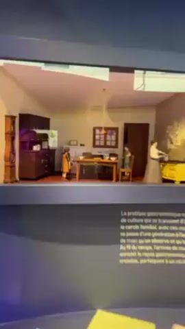 The video shows two framed dioramas, depicting kitchen scenes with miniature figures. The display is part of an exhibit, possibly about food culture or traditions. Text panels in French and English are visible beneath each diorama, further explaining the exhibit's themes. The dioramas are illuminated, creating depth and drawing attention to the miniature scenes.