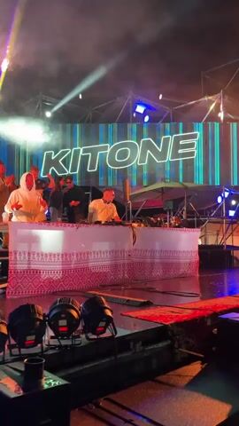 A group of people are on a stage in front of a large LED screen that displays the word "KITONE". The screen has a blue and green striped background. A DJ is at the center of the stage, operating turntables and mixers. The DJ is wearing sunglasses and a white t-shirt. Two other individuals are standing behind the DJ, one holding a drink and the other wearing a hooded jacket. Several stage lights are positioned in the foreground, and a patterned fabric drapes over the DJ booth. The overall atmosphere appears to be that of a live music performance or event.