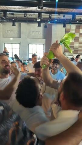 A group of people are gathered in a room, possibly a venue or hall, with a stage and band visible in the background. Several individuals, mostly men wearing white t-shirts, are in the foreground, surrounding one person who is holding up a large, bright yellow and green fan. The person with the fan is in the process of pouring a drink from a bottle into the open mouth of another person in the group. The atmosphere appears celebratory, with people smiling and engaged in the activity. The room has wooden accents on the ceiling and walls, and there are windows visible in the background.
