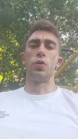 The media file shows a close-up, selfie-style video of a young man outdoors, with a focus on his face and upper body. He is wearing a white sleeveless t-shirt with a black logo that reads "THE NORTH FACE". He has short, light brown hair and appears to be in his late twenties or early thirties. His expression is somewhat intense, and his mouth is slightly open, suggesting he might be speaking or about to speak. The background is blurred but shows lush green foliage, indicative of a park or wooded area. Sunlight filters through the leaves, suggesting it is daytime. There are hints of outdoor gym equipment in the background, with yellow and dark metal structures visible. The overall impression is that the man is taking a break during or after an outdoor workout. The context provided places the scene in Fastiv, Ukraine.